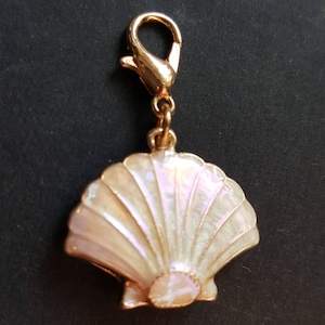 May include: A gold-colored charm with a pearlescent seashell design. The charm features a hinged clasp for easy attachment to a bracelet or necklace. The seashell has a detailed, fan-like pattern.