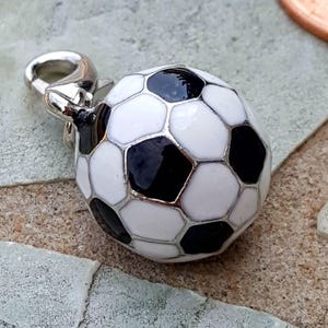 Pode incluir: Um berloque de bola de futebol com fecho prateado. O berloque é branco com pentágonos pretos, delineados em prata. O berloque é uma réplica em miniatura de uma bola de futebol.