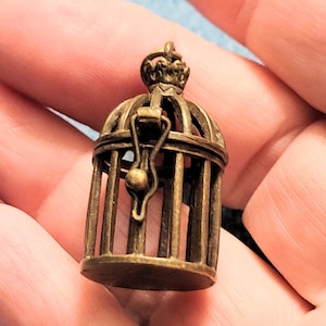May include: A small, antique-looking, bronze birdcage charm with a crown on top. The cage has a hinged door and a small ball hanging inside.