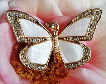 Puede incluir: Un colgante de mariposa dorado con alas de nácar blanco y brillantes transparentes. El colgante está sobre una rosa rosa.