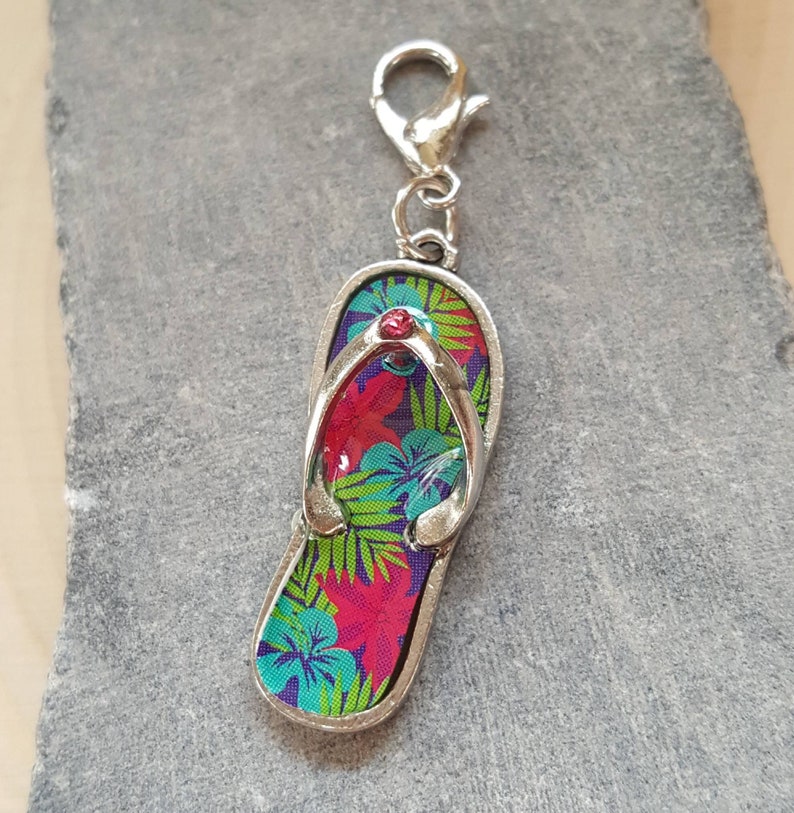 May include: A silver charm in the shape of a flip-flop with a tropical floral print in shades of pink, green, and blue. The flip-flop has a red rhinestone in the center.