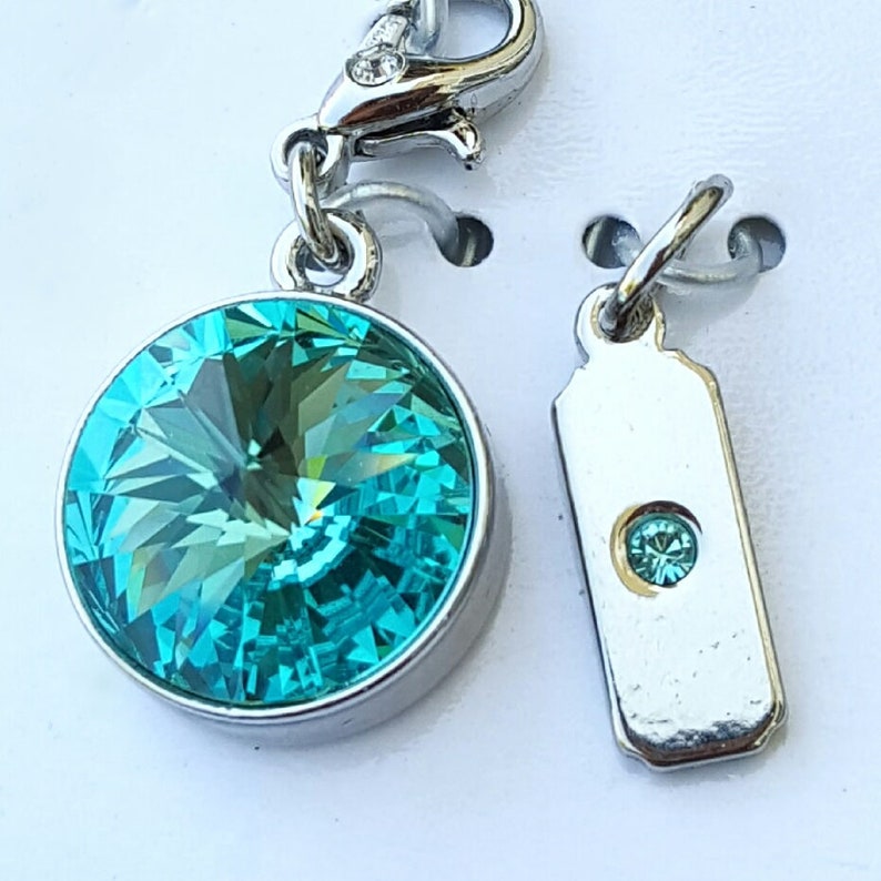 May include: A silver charm featuring a turquoise blue crystal, and a smaller silver charm with a turquoise blue crystal. The charms are attached to a silver chain.