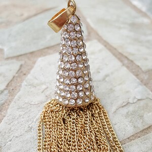 May include: A gold-toned pendant with a cone-shaped top encrusted with clear rhinestones. Below the cone, a cascade of gold chains forms a tassel. The pendant has a small loop at the top for hanging.