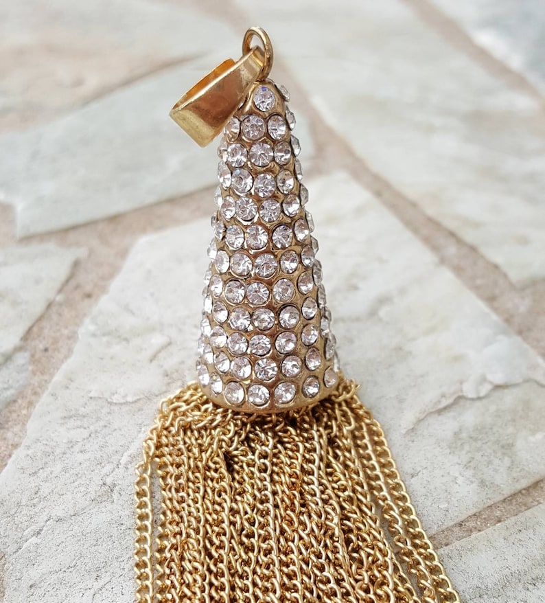 May include: A gold-toned pendant with a cone-shaped top encrusted with clear rhinestones. Below the cone, a cascade of gold chains forms a tassel. The pendant has a small loop at the top for hanging.