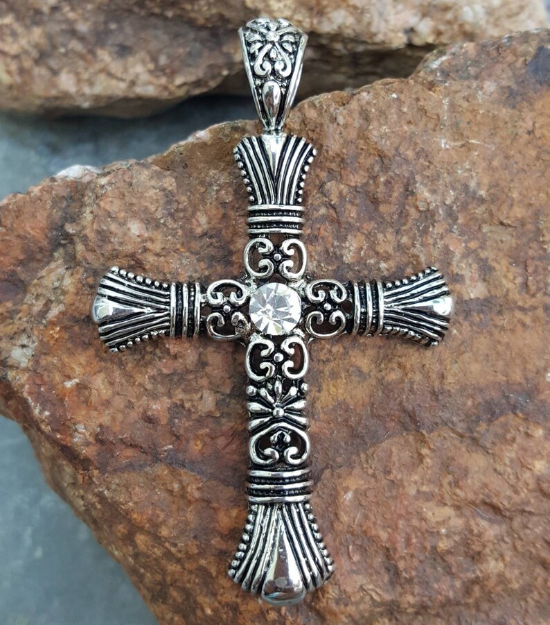 May include: A silver cross pendant with an ornate design and a clear rhinestone in the centre.