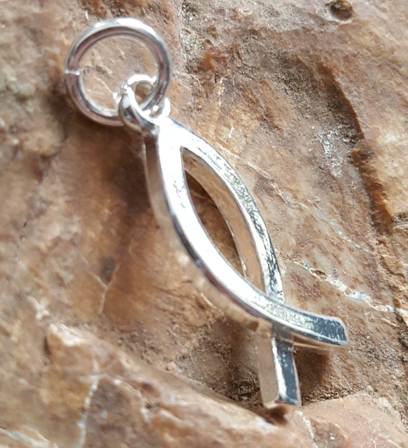May include: Silver charm in the shape of a stylized fish with a cross at the bottom. The charm has a loop at the top for attaching to a necklace or bracelet.