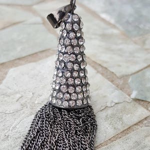 May include: A silver-toned pendant with a cone-shaped top encrusted with clear crystals. Below the crystal-covered section, a cascade of silver chains forms a tassel. The pendant has a small loop at the top for hanging.