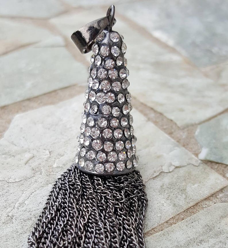 May include: A silver-toned pendant with a cone-shaped top encrusted with clear crystals. Below the crystal-covered section, a cascade of silver chains forms a tassel. The pendant has a small loop at the top for hanging.