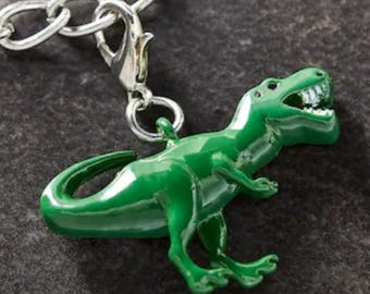 May include: A shiny green Tyrannosaurus Rex charm with a silver-colored clasp and chain. The dinosaur has a detailed, open mouth with visible teeth. The charm is attached to a silver chain, suitable for a bracelet or necklace.
