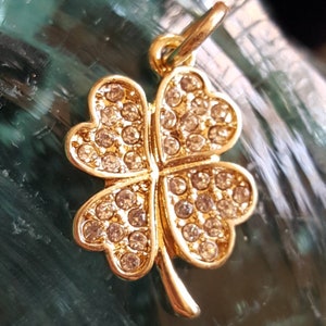 May include: A gold four-leaf clover charm with clear rhinestones. The charm has a loop at the top for attaching to a necklace or bracelet.