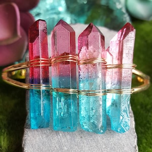 Può includere: Un braccialetto avvolto in filo dorato con cinque punte di cristallo rosa e blu sfumate. I cristalli sono disposti verticalmente e sono fissati con filo dorato.