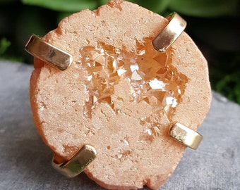 May include: A peach-colored geode with a large crystal cavity filled with clear quartz crystals. The geode is surrounded by four gold metal bands.