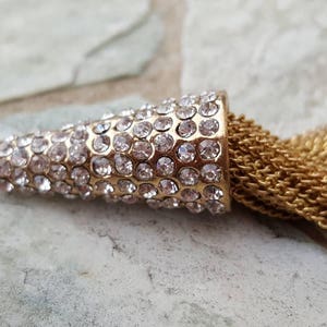 May include: A gold-colored pendant with a cone-shaped top encrusted with clear rhinestones. A cascade of gold chain tassels hangs from the pendant. The pendant is lying on a light gray stone surface.