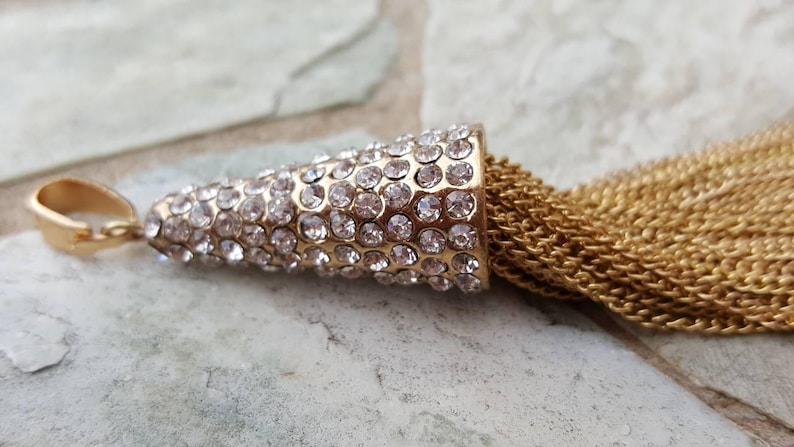 May include: A gold-colored pendant with a cone-shaped top encrusted with clear rhinestones. A cascade of gold chain tassels hangs from the pendant. The pendant is lying on a light gray stone surface.