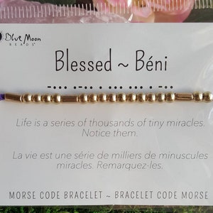 Puede incluir: Pulsera de cuentas doradas con un cordón morado y una tarjeta que dice "Blessed ~ Béni" en texto negro. La tarjeta también incluye el código Morse para la palabra "Blessed" y la frase "Life is a series of thousands of tiny miracles. Notice them." en inglés y francés.
