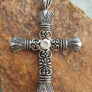 May include: A silver ornate cross pendant with a clear rhinestone in the centre. The cross has a detailed design with intricate scrollwork and a bail at the top.