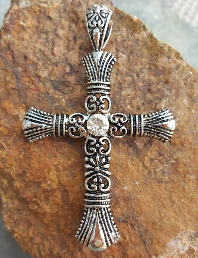 May include: A silver ornate cross pendant with a clear rhinestone in the centre. The cross has a detailed design with intricate scrollwork and a bail at the top.