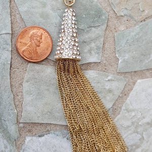 May include: A gold-colored tassel pendant with a cone-shaped top encrusted with clear rhinestones. The tassel is made of multiple strands of gold-colored chain. A penny is next to the pendant for size comparison.