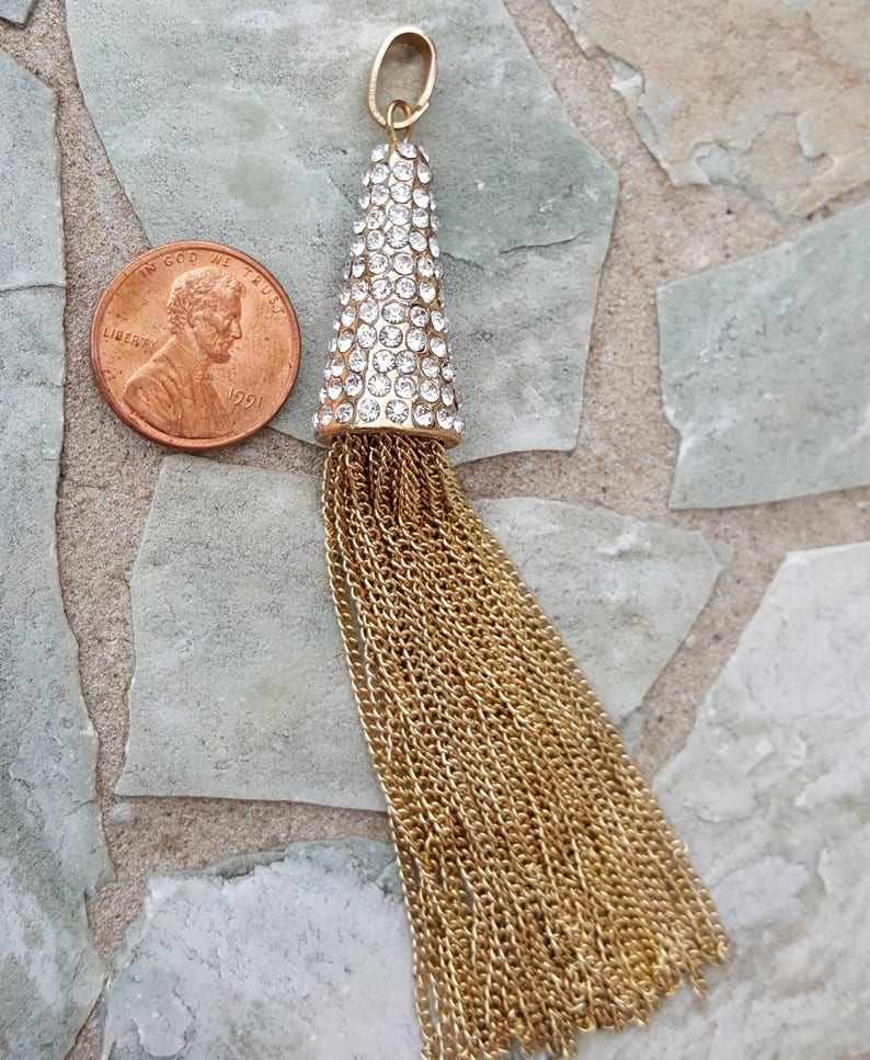 May include: A gold-colored tassel pendant with a cone-shaped top encrusted with clear rhinestones. The tassel is made of multiple strands of gold-colored chain. A penny is next to the pendant for size comparison.