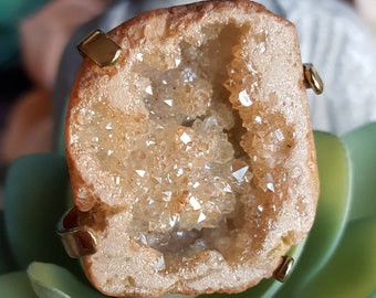 May include: A geode ring with a gold setting. The geode is filled with clear quartz crystals and has a brown exterior.
