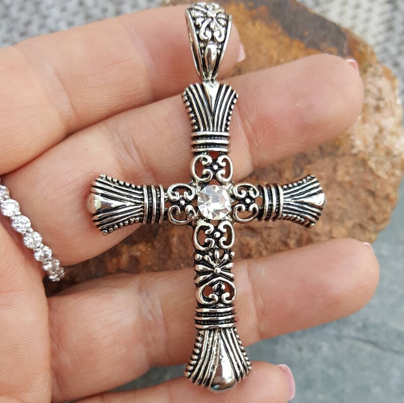 May include: A silver cross pendant with an ornate design and a clear rhinestone in the centre.