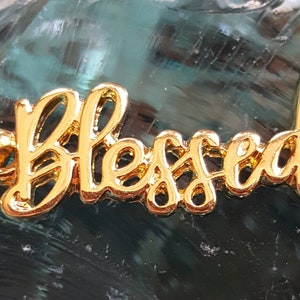 May include: Gold-colored metal charm with the word "Blessed" in a cursive font.