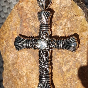 May include: A silver cross pendant with an ornate design and a clear rhinestone in the centre.