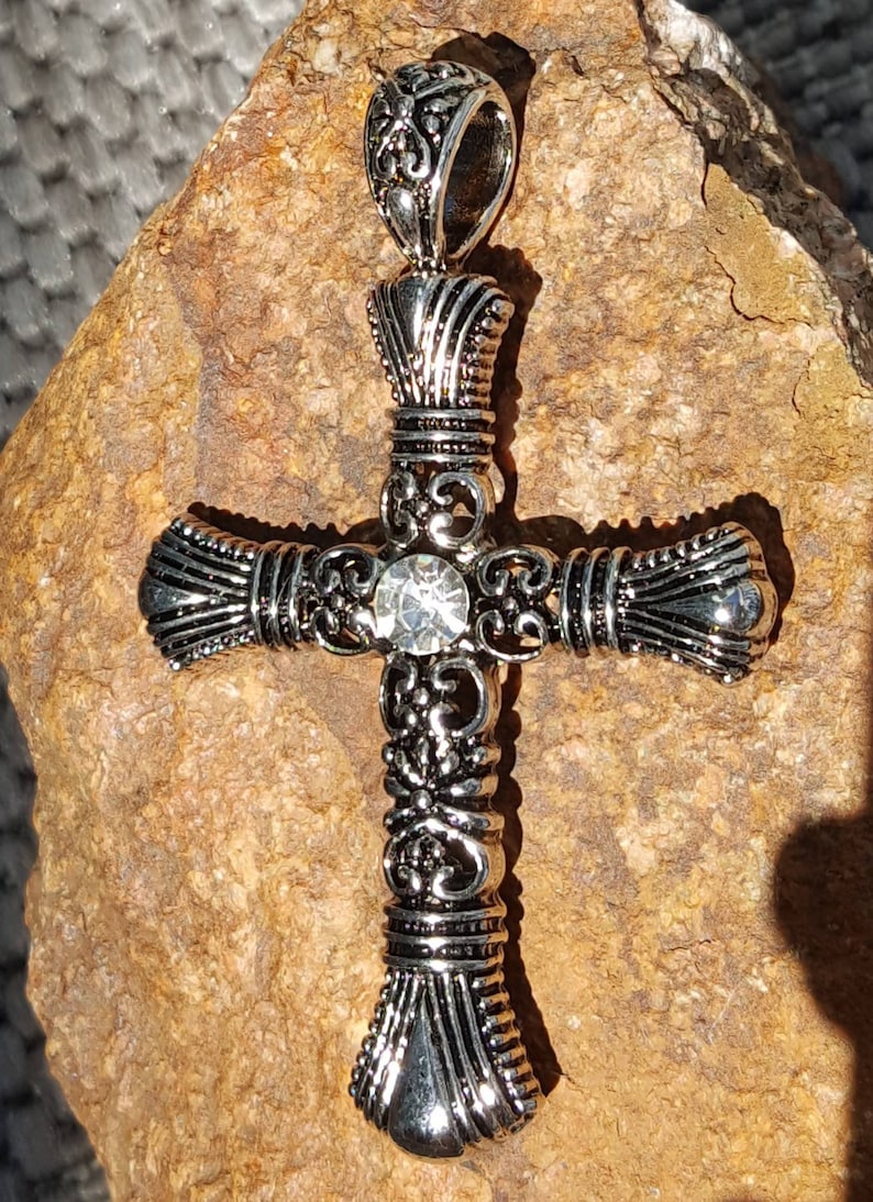 May include: A silver cross pendant with an ornate design and a clear rhinestone in the centre.