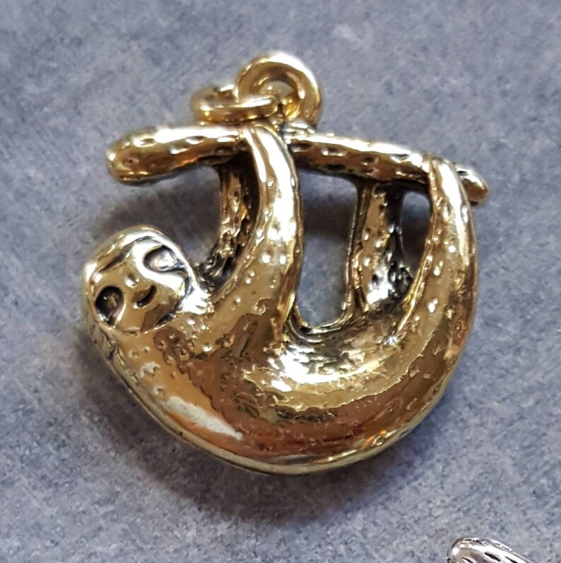K&ouml;nnte beinhalten: Goldfarbener Metallanh&auml;nger eines Faultiers, das an einem Ast h&auml;ngt. Der Anh&auml;nger ist detailliert mit einer strukturierten Oberfl&auml;che und einem l&auml;chelnden Gesicht.