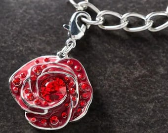 Puede incluir: Una pulsera con colgante plateada con un colgante de rosa roja. El colgante de rosa está detallado con esmalte rojo y pequeños cristales rojos, y tiene un gran cristal rojo en el centro. La pulsera está sobre un fondo oscuro.