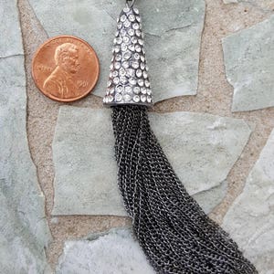 May include: A silver-toned pendant with a cone-shaped top encrusted with clear crystals and a long chain tassel. The pendant is shown next to a U.S. penny for size comparison. The background is a stone surface.