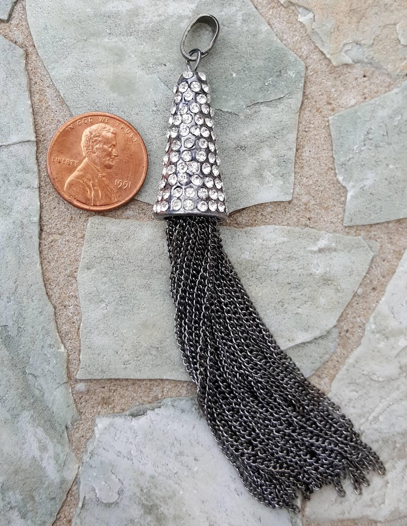 May include: A silver-toned pendant with a cone-shaped top encrusted with clear crystals and a long chain tassel. The pendant is shown next to a U.S. penny for size comparison. The background is a stone surface.