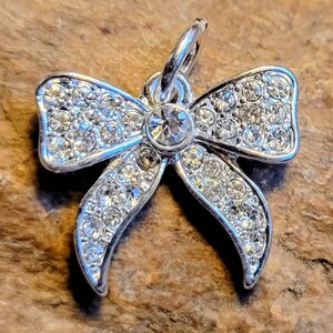 May include: A silver charm in the shape of a bow with a clear crystal in the center. The bow is covered in small clear crystals.