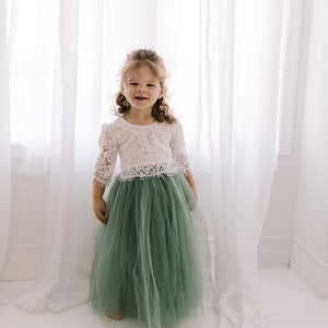 May include: A young girl wearing a white lace top and a sage green tulle skirt. She is standing in front of a white curtain and smiling.