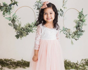Puede incluir: Un vestido largo hasta el suelo con un corpiño de encaje blanco de manga tres cuartos y una falda amplia de tul rosa pálido en capas. La prenda se combina con un accesorio de lazo para el pelo con lentejuelas oro rosa.