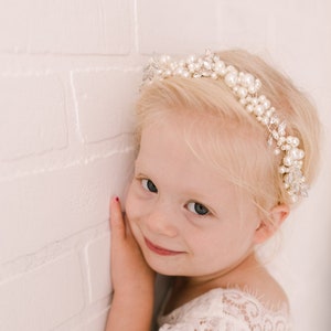 May include: A young girl with blonde hair wearing a white lace dress and a pearl and rhinestone headband. She is smiling and looking at the camera.