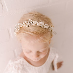 May include: A young girl wearing a white dress with a pearl and crystal hairpiece. The hairpiece is a delicate headband with small pearls and rhinestones.