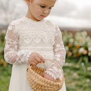 May include: A young girl in a white lace dress holds a woven basket. She is wearing a flower crown and looking down at the basket.