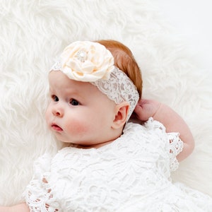 K&ouml;nnte beinhalten: Ein Baby M&auml;dchen in einem wei&szlig;en Spitzenkleid und einem Haarreif mit Pfirsichbl&uuml;ten liegt auf einem wei&szlig;en, flauschigen Teppich.