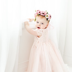 May include: A pale pink dress with a lace bodice and a layered tulle skirt, worn with a floral crown featuring pink and white flowers. The dress has short lace sleeves and a full, flowing skirt that reaches the floor, creating a soft appearance.