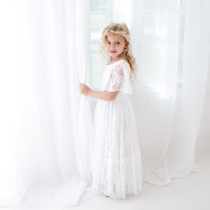 May include: A young girl wearing a white lace dress with a gold crown, standing by a white curtain and looking at the camera.