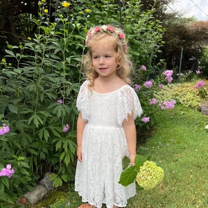 May include: A young girl wearing a white lace dress with a floral crown and holding a bouquet of white flowers. She is standing in a grassy area with green foliage and flowers.