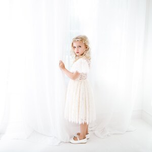 May include: A cream-colored lace dress with short sleeves and a scalloped hemline, paired with a gold-toned floral headpiece and white flat shoes. The garment features intricate lace detailing on the bodice and skirt, creating a textured appearance.
