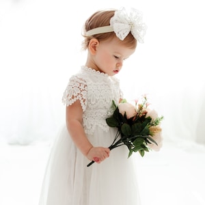 May include: A child in a white lace dress and a matching headband with a large bow. The child holds a bouquet of pink and cream flowers with green leaves. The dress has a tulle skirt, creating a soft, flowing look.