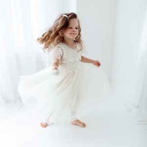 May include: A young girl twirls in a cream-colored dress with a lace bodice and flowing tulle skirt. Her long, wavy hair is adorned with a small hair accessory. The dress is paired with white leggings. The background is a bright white space.