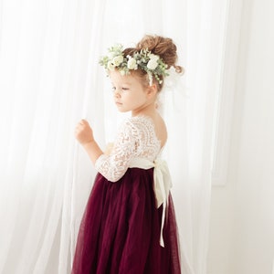 May include: A young girl wearing a white lace bodice dress with a burgundy tulle skirt. She is wearing a flower crown and a white sash with a bow. The dress has a low back.