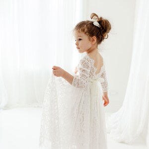May include: A floor-length white lace dress featuring three-quarter length sleeves, a scalloped neckline, and a deep scoop back. The garment includes a solid white ribbon tied in a bow at the waist, with intricate floral lace patterns throughout.