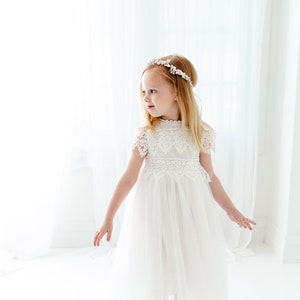 May include: A white dress featuring a lace bodice with short sleeves and a high neckline, paired with a layered tulle skirt. The garment is accessorized with a delicate floral headband featuring small white flowers and beads.