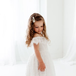 May include: A young girl wearing a white off-the-shoulder lace dress with a high-low hemline. She has curly brown hair and is wearing a pearl hair clip.