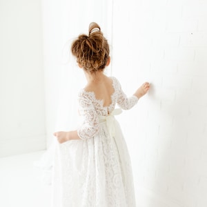 May include: A white lace dress with three-quarter length sleeves, a scalloped scoop neckline, and a floor-length skirt. The garment features a fitted bodice with a sheer lace back and a solid white ribbon tied into a bow at the waistline.